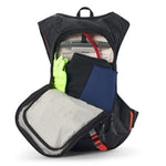USWE Moto Hydro 12L hydration pack fully opened to reveal dual-compartment interior with 3.0L hydration bladder in upper section, neon yellow gloves tucked in side pocket, folded navy blue and black clothing layers in main storage area, and gray jacket in lower compartment with red pull tabs on zippers.