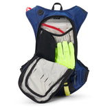 USWE Moto Hydro 8L hydration pack with both compartments unzipped showing 3.0L Elite Hydration Bladder with red hose, yellow-green riding gloves, gray clothing layer, and black mesh storage divider inside the main compartment for dirt bike gear organization.