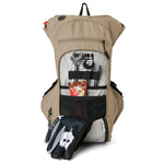 USWE OUTLANDER Moto 9L hydration pack in Sand colorway displayed vertically with front tool pocket unzipped exposing chrome wrenches, emergency contact card, and energy bar secured by black elastic straps, plus dirt bike gloves positioned at base of pack.